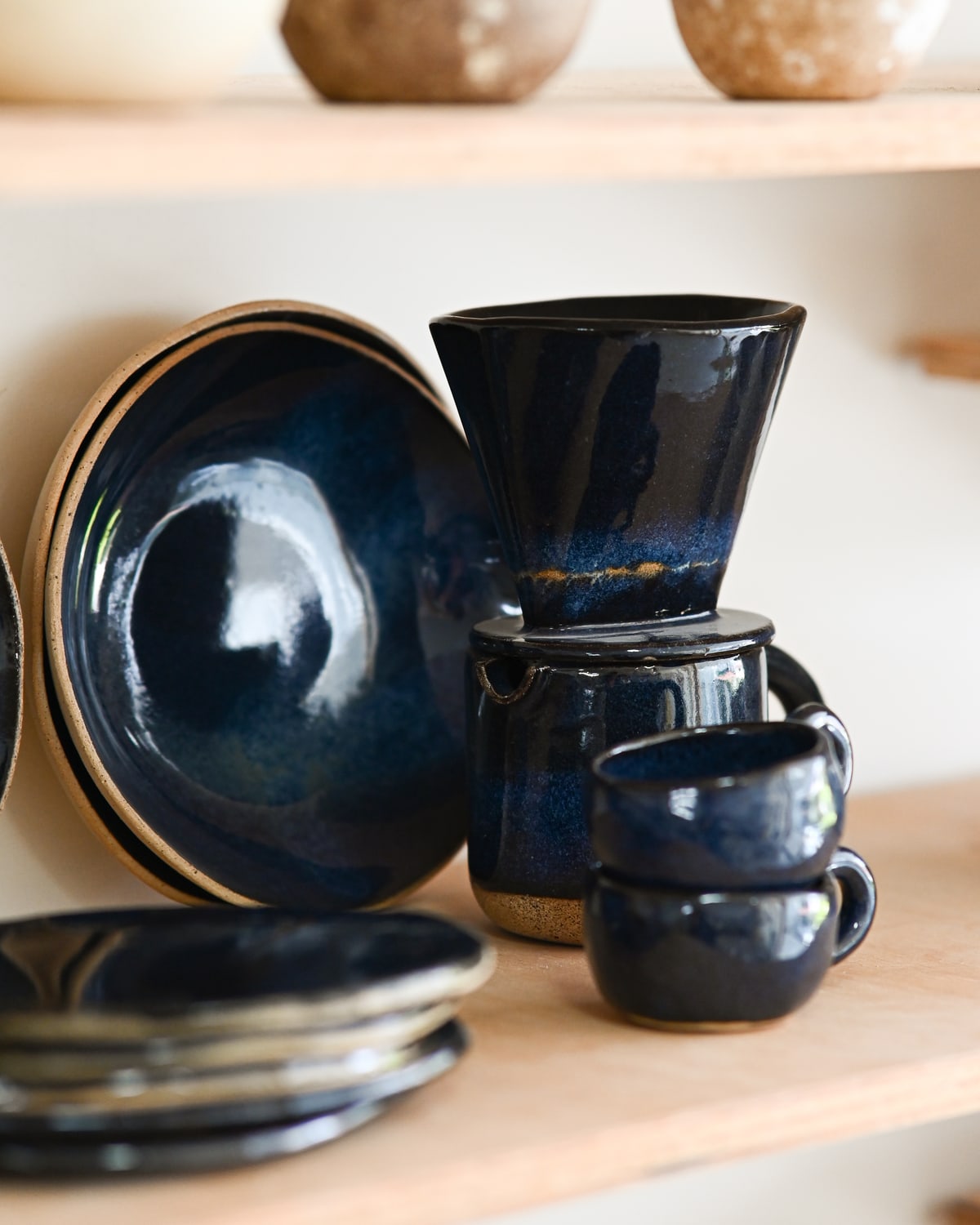 Um jogo de café de cerâmica esmaltada em azul profundo na prateleira do ateliê: jarra, coador, duas xícaras e pratos empilhados