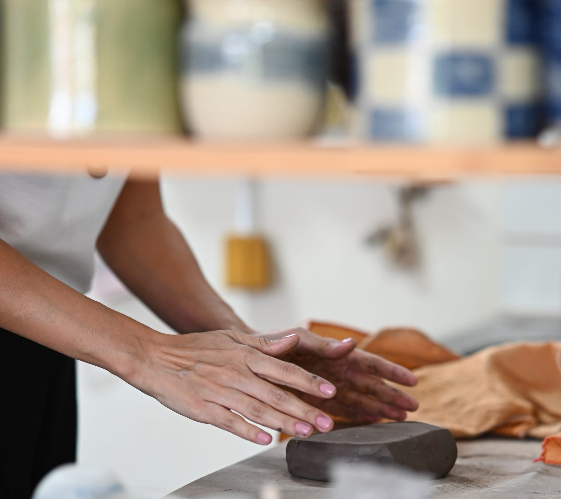 Duas mãos alisando um bloco de argila sobre a mesa de trabalho do ateliê