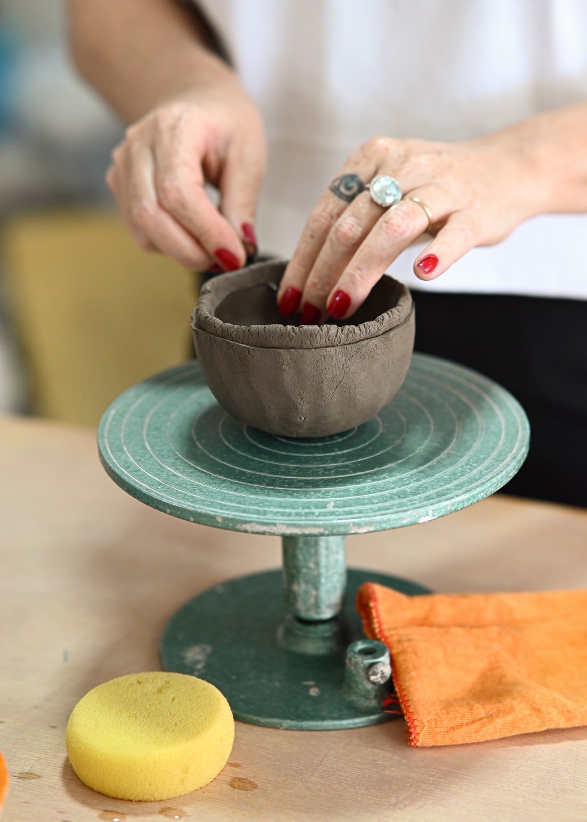 Duas mãos modelando uma cumbuca de argila sobre um torno de mão verde, com a esponja e o pano ao lado