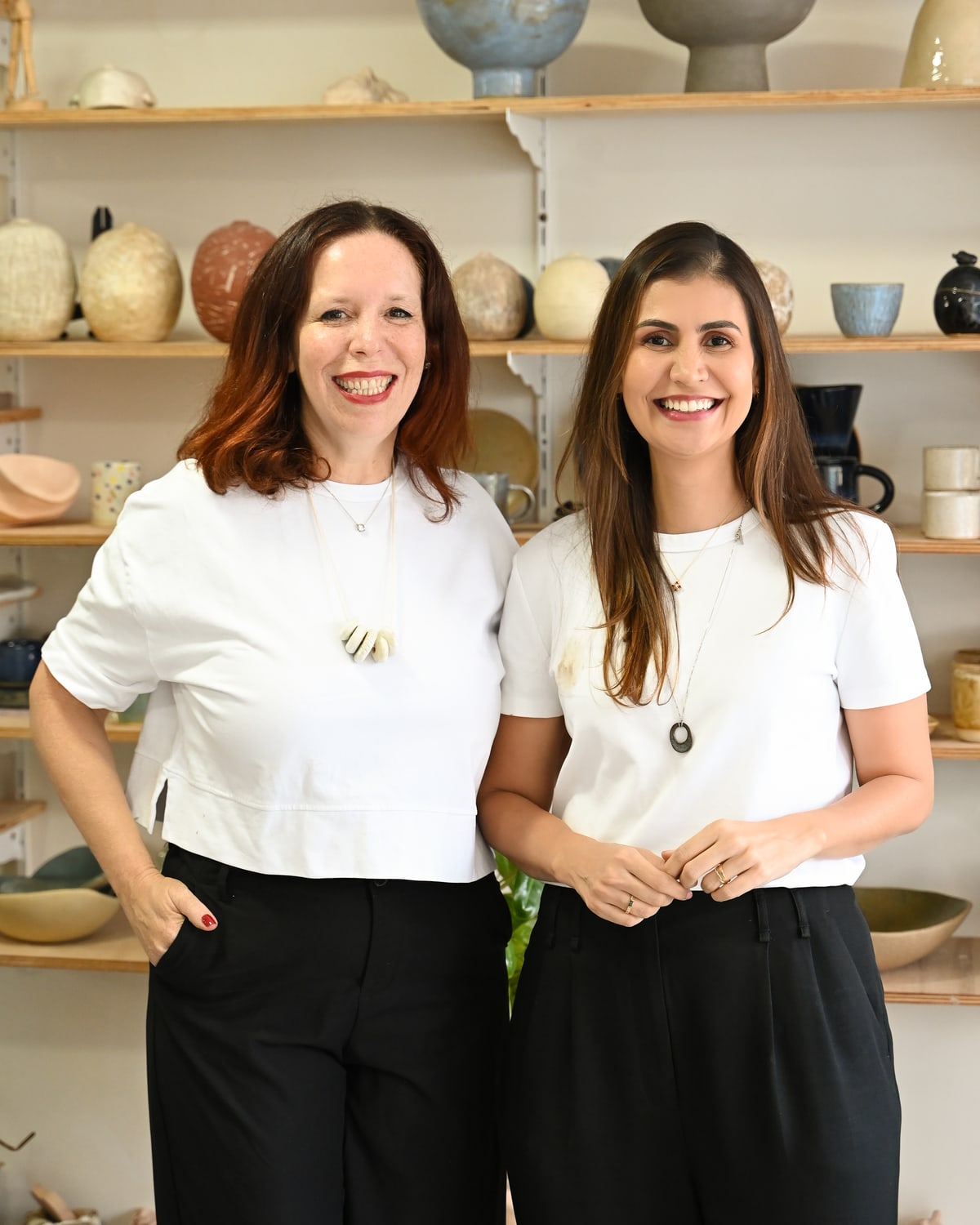Isa e Marcela de pé no ateliê, lado a lado e sorrindo, com as prateleiras de peças atrás