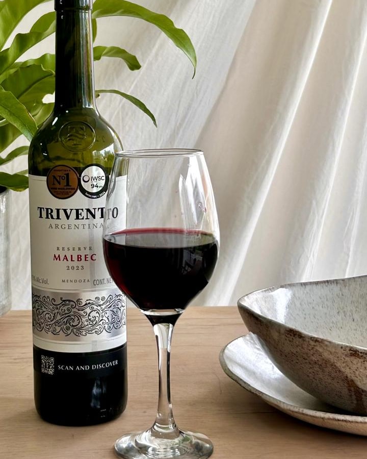 Uma garrafa de vinho tinto e uma taça servida sobre a mesa de madeira do ateliê, ao lado de uma cumbuca e um prato de cerâmica